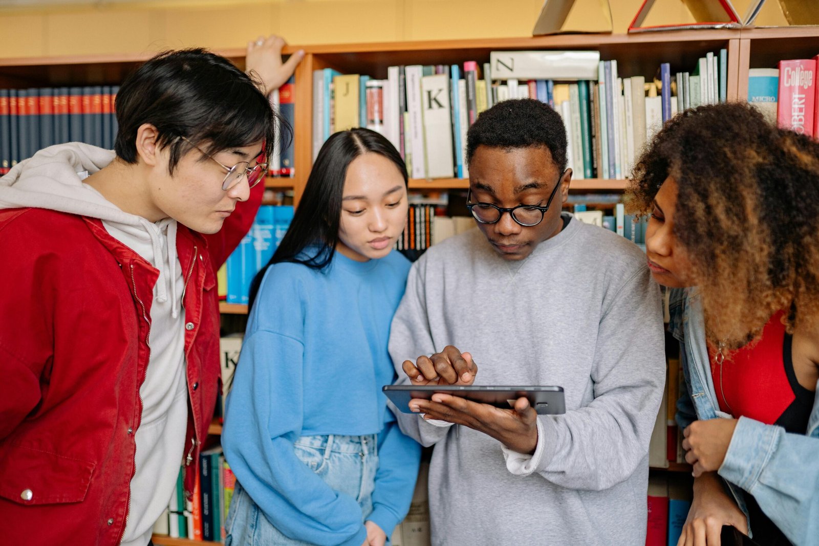 A group of college students collaborate on a tablet in a library surrounded by bookshelves.